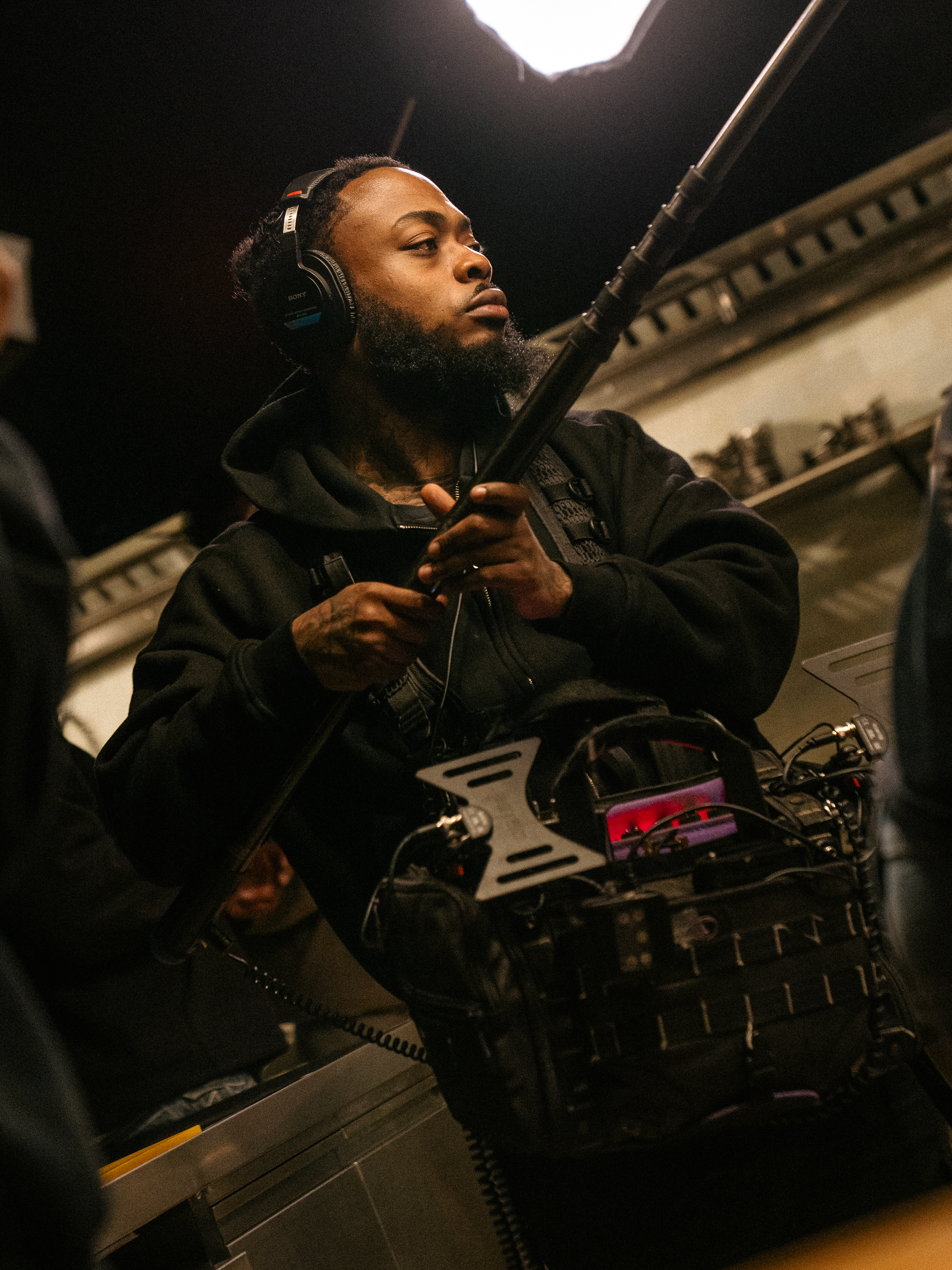 Sound mixer on location holding a boom pole, headphones on, recorder rig in front